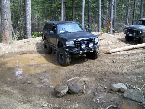 1991 Ford Bronco - A modified black 1992 Ford Bronco navigates a muddy forest trail, with another vehicle visible in the background.