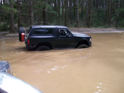 1991 Ford Bronco - A black Ford Bronco navigates through a muddy water obstacle in a wooded area.