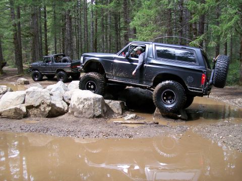 1991 Ford Bronco - Two Ford Broncos tackle a challenging off-road course with muddy puddles and large rocks.