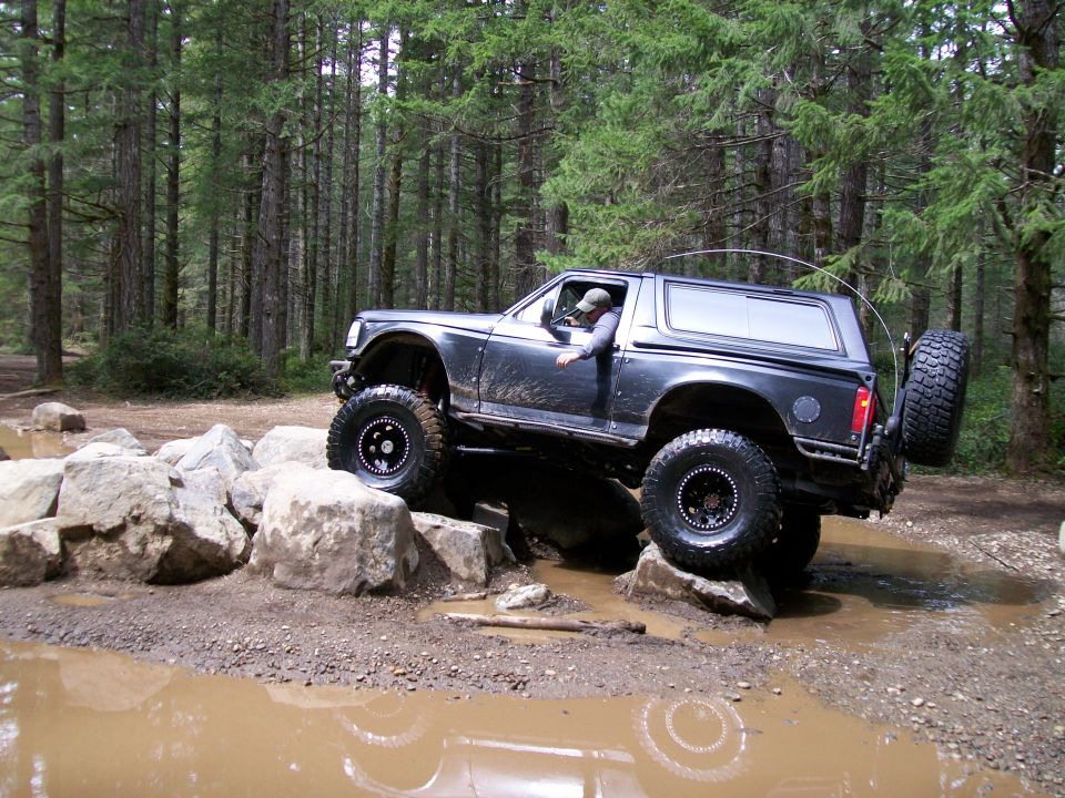 Dark Ford Bronco navigating a rocky, muddy trail in a forest. Driver is visible