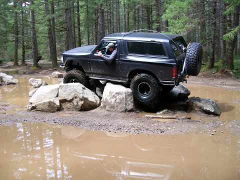 1991 Ford Bronco - A black 1991 Ford Bronco tackles an off-road obstacle course, carefully maneuvering over large rocks and through muddy water.