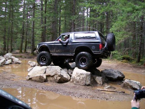 1991 Ford Bronco - A black Ford Bronco navigates a rocky obstacle on a wooded trail, with a person filming from the side.