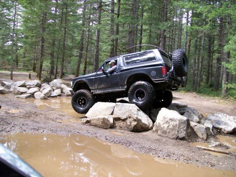 1991 Ford Bronco - A modified SUV tackles a challenging off-road trail with large rocks and mud.