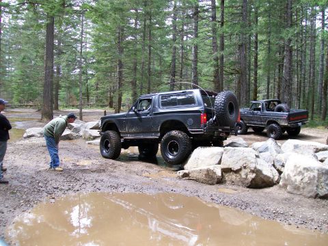 1991 Ford Bronco - Two Ford off-road vehicles, a Bronco and a pickup, tackle a challenging muddy course in a forest.