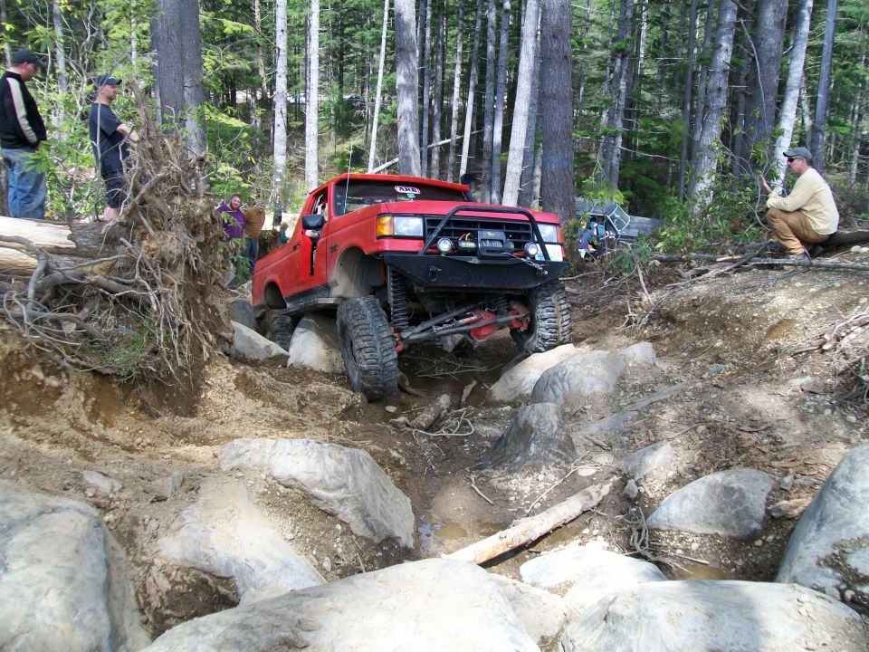 Red 1990s Ford F-Series truck off-roading over large rocks. Another vehicle is visible in the background