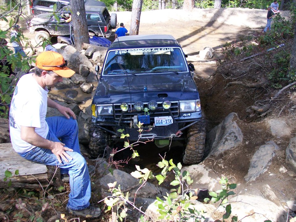 1991 Ford Bronco navigating a steep, rocky trail with a man sitting nearby. Another Bronco is visible in the background