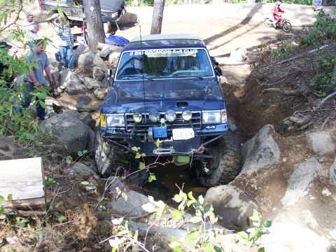 1991 Ford Bronco - A modified Ford Bronco attempts a challenging obstacle on an off-road course, with other vehicles and spectators visible.