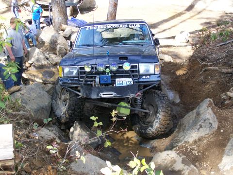 1991 Ford Bronco - A dark blue Ford Bronco tackles a challenging off-road course with large rocks and mud.