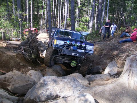 1991 Ford Bronco - A blue 1991 Ford Bronco tackles a challenging rocky obstacle on an off-road trail, with other vehicles and spectators in the background.