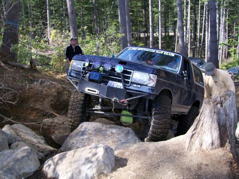 1991 Ford Bronco - A modified Ford Bronco tackles a challenging off-road obstacle in a wooded area.