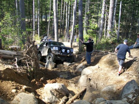 1991 Ford Bronco - An off-road SUV navigates a challenging, muddy, and rocky trail through a dense forest with spectators watching.
