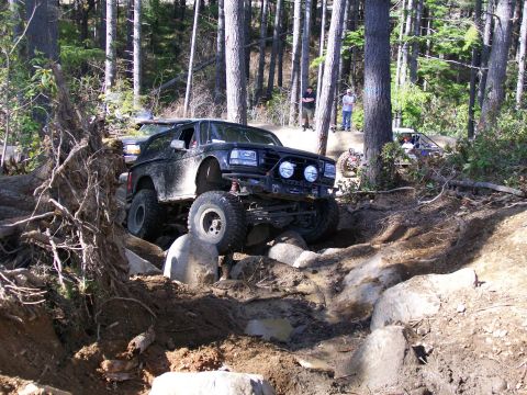 1991 Ford Bronco - A modified black SUV tackles a challenging off-road course in a forest setting.