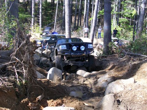 1991 Ford Bronco - A modified black Ford Bronco tackles a challenging rocky obstacle on a wooded trail, with other vehicles and spectators nearby.