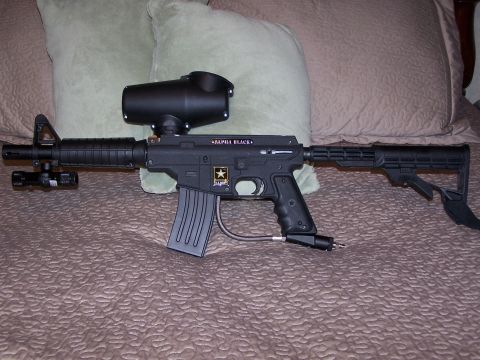 1986 Ford Bronco - A black paintball gun with an attached hopper and adjustable stock rests on a bed.