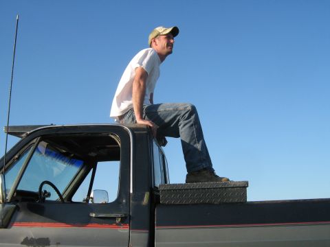 1990 Ford Bronco - Man sits atop a pickup truck cab against a clear blue sky. The truck appears to be from the 1980s.