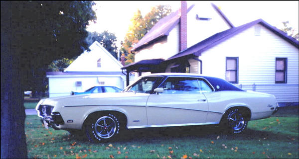 Side view of white 1969 Mercury Cougar with black vinyl roof parked on grass in front of houses