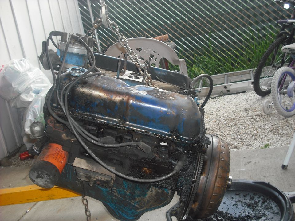 Engine on a yellow engine hoist, covered in oil. A chain is attached to it. There's a white plastic chair and a ladder in the background