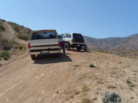 1995 Ford Bronco - Two SUVs are parked on a dirt incline under a clear blue sky, with people attending to the white vehicle.