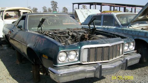 1972 Ford Ranchero - A dark green 1972 Ford Ranchero is parked in a junkyard with its hood open, revealing the engine. Another light blue vehicle is parked clos…