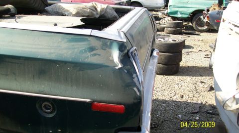 1972 Ford Ranchero - A close-up rear-side view of a damaged dark green vehicle, likely a pickup truck or El Camino style, in a junkyard setting. The vehicle has…