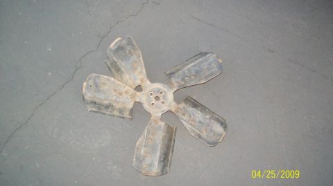 1972 Ford Ranchero - A metal fan with five blades is shown from a top-down perspective, lying on a grey, cracked concrete surface. The fan appears to be a vehic…