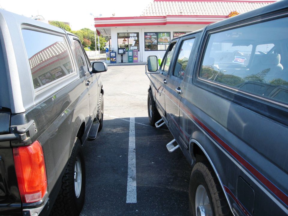 Two Ford Broncos parked in a lot, one dark gray, one black. Building in background