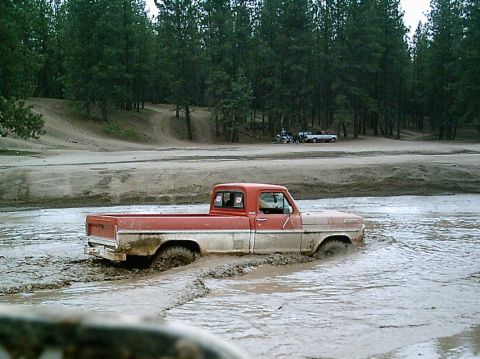 1968 Ford F-250 - A two-tone pickup truck navigates through a muddy terrain with trees in the background.