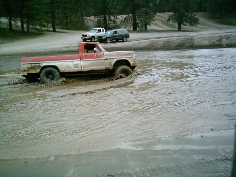 1968 Ford F-250 - A red and white Ford F-250 truck navigates through a muddy off-road course with other vehicles nearby.