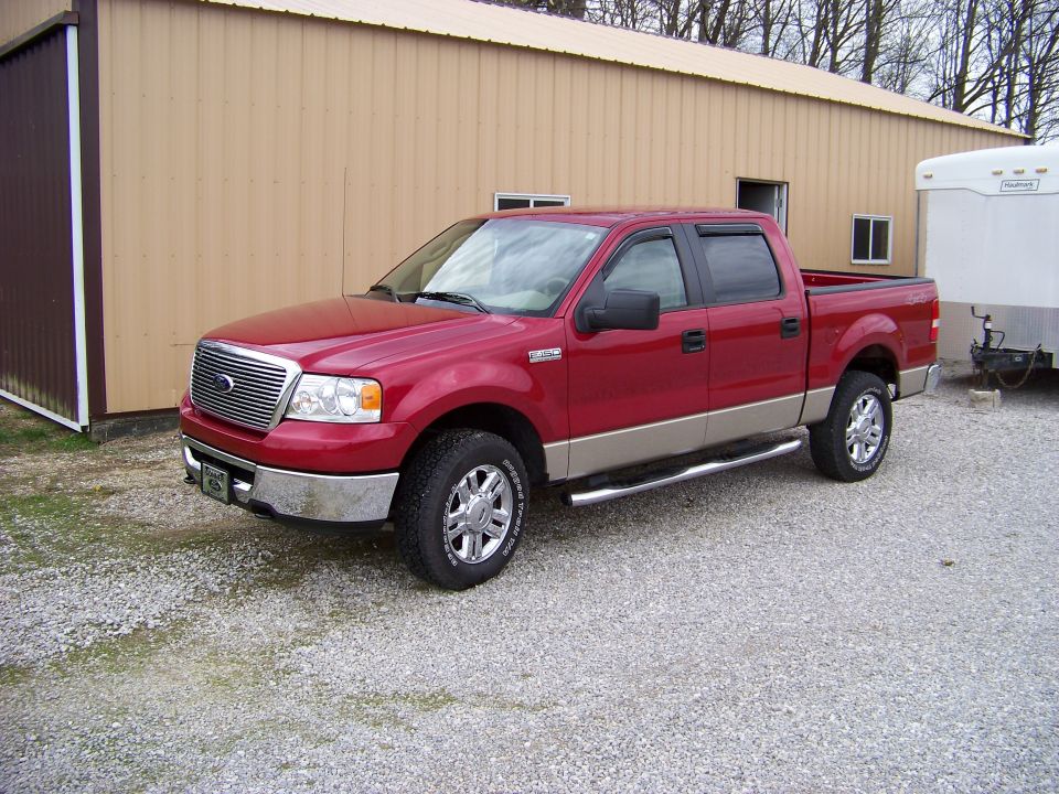 Red and tan Ford F-150 SuperCrew pickup parked on gravel by a tan pole barn