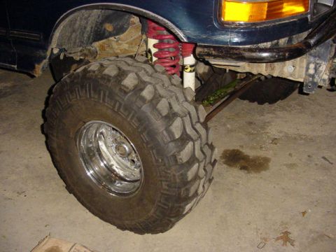 1992 Ford Bronco - Front wheel close-up of a lifted dark blue Ford truck in a garage, showing an oversized square-lug mud tire on a polished chrome wheel. A r…