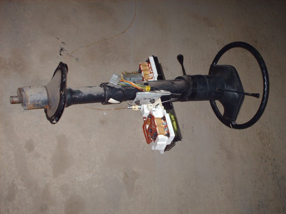 Removed black steering column with steering wheel, wiring connectors and switches lying on concrete floor