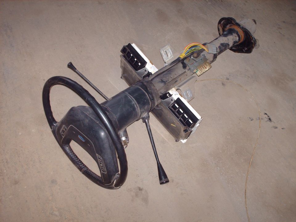 Removed steering column with black steering wheel and wiring harness lying on concrete floor