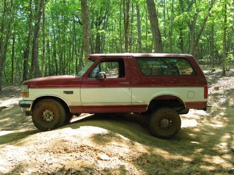 1995 Ford Bronco - A two-tone red and white Ford Bronco is seen from the side, parked on a dirt path surrounded by trees.