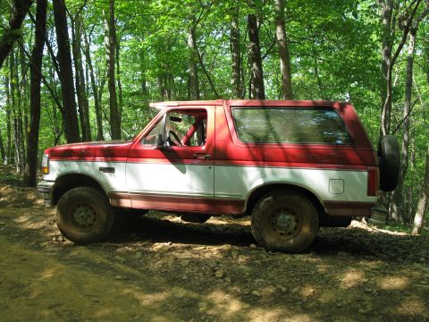 1995 Ford Bronco - A two-tone red and white Ford Bronco is seen from the side on a wooded trail.