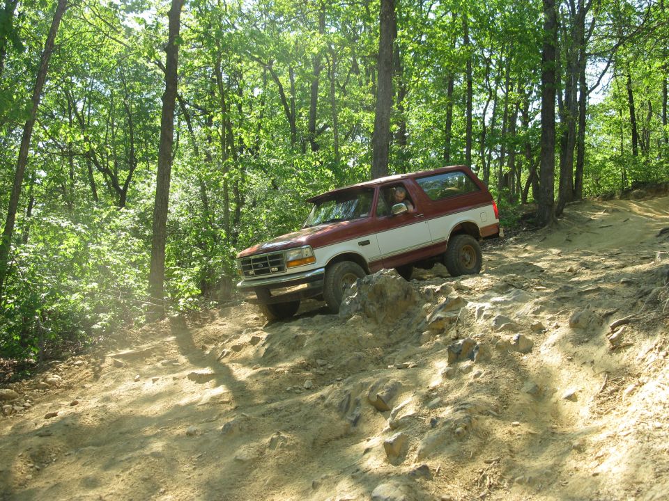 1995 Ford Bronco driving on a rocky off-road trail in a forest.