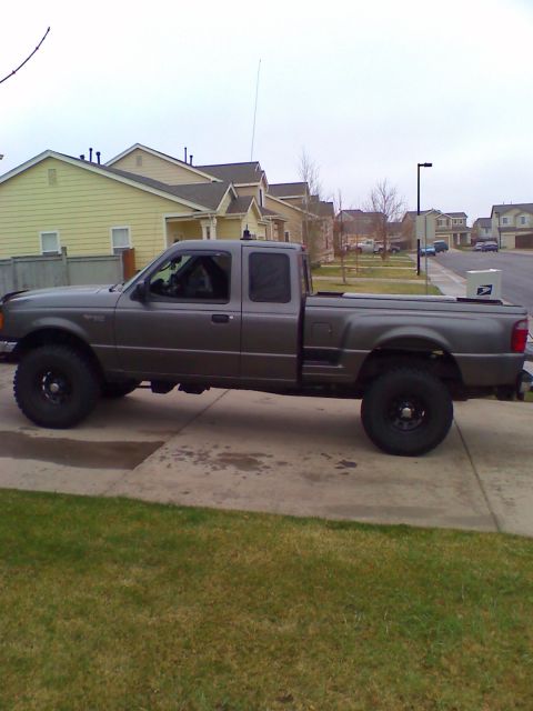 2004 Ford Ranger - Side profile of a gray Ford Ranger extended-cab pickup with a lift kit, black steel wheels and large off-road tires, parked in a concrete d…