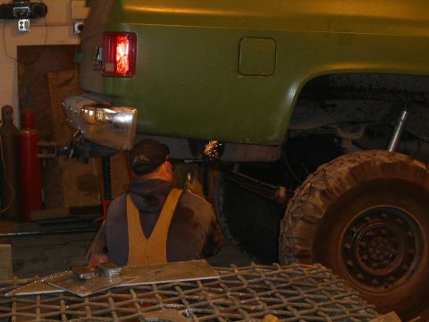 1990 Chevrolet Suburban - A person wearing a welding mask and a tan apron is working on the rear bumper of a green vehicle, possibly a Chevrolet Suburban. The vehicl…