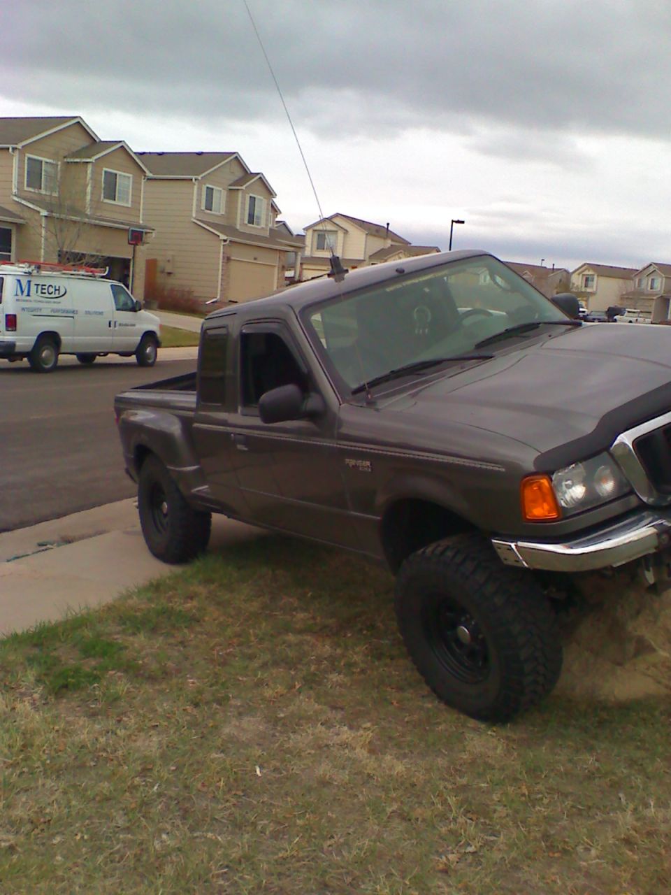Dark gray Ford Ranger pickup truck parked on grass with houses and a white work van in background.