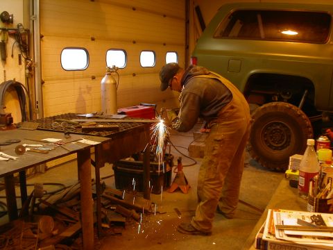 1990 Chevrolet Suburban - A person in overalls is welding in a garage next to a green Chevrolet Suburban. Sparks are flying from the welding torch as the person work…