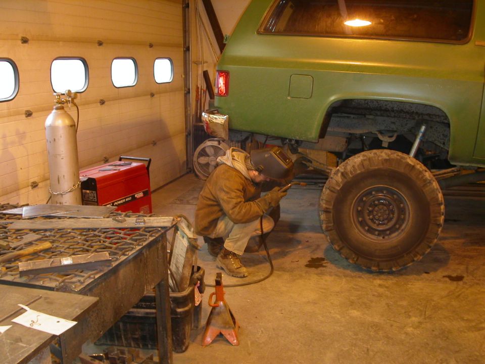 Green Chevrolet Suburban being welded under the rear quarter panel with a large tire visible.