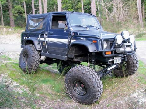 1989 Ford Bronco - A dark blue Suzuki Samurai-style soft-top convertible SUV lifted on large off-road mud tires, shown from the front passenger side with a ro…