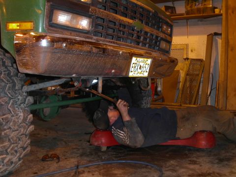 1990 Chevrolet Suburban - A person lies on a creeper underneath the front of a green Chevrolet pickup truck, working on the suspension with a wrench. The truck has l…