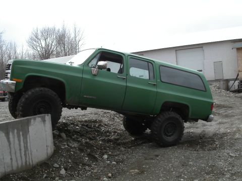 1990 Chevrolet Suburban - A lifted green Chevrolet Suburban is shown at an angle, with its front tire on top of a concrete barrier and its rear tire kicking up dust.…