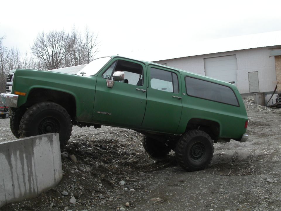 Green lifted Chevrolet Suburban off-roading over a concrete barrier.