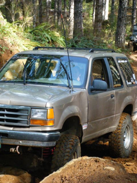 2005 Jeep Wrangler - A tan Ford Explorer is shown from the front 3/4 angle, navigating an off-road trail with large mud tires. The vehicle is surrounded by tree…