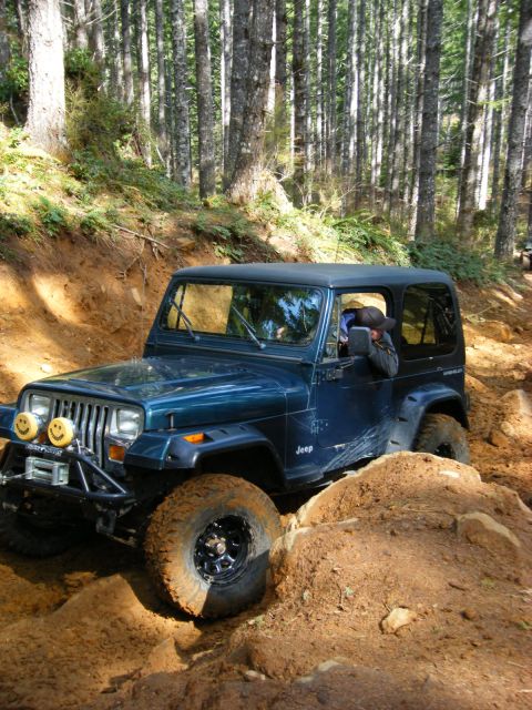 2005 Jeep Wrangler - A dark blue Jeep Wrangler is navigating a steep, muddy trail in a forest. The vehicle is angled to the right, with its front tire on the le…