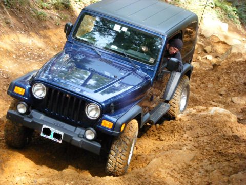 2005 Jeep Wrangler - A blue Jeep Wrangler is navigating a steep, muddy trail with large rocks. The driver, wearing a black beanie, is visible through the open d…