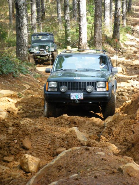 2005 Jeep Wrangler - Two Jeep SUVs are navigating a muddy, rocky trail through a forest. The front Jeep is dark green and appears to be a Cherokee XJ model, whi…