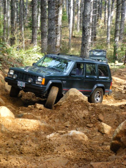 2005 Jeep Wrangler - A dark green Jeep Cherokee is navigating a steep, rocky, and muddy trail in a forest. Another Jeep is visible in the background.