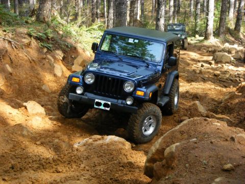 2005 Jeep Wrangler - A dark blue Jeep Wrangler is navigating a rocky, muddy trail in a forest. Another Jeep is visible further up the trail.